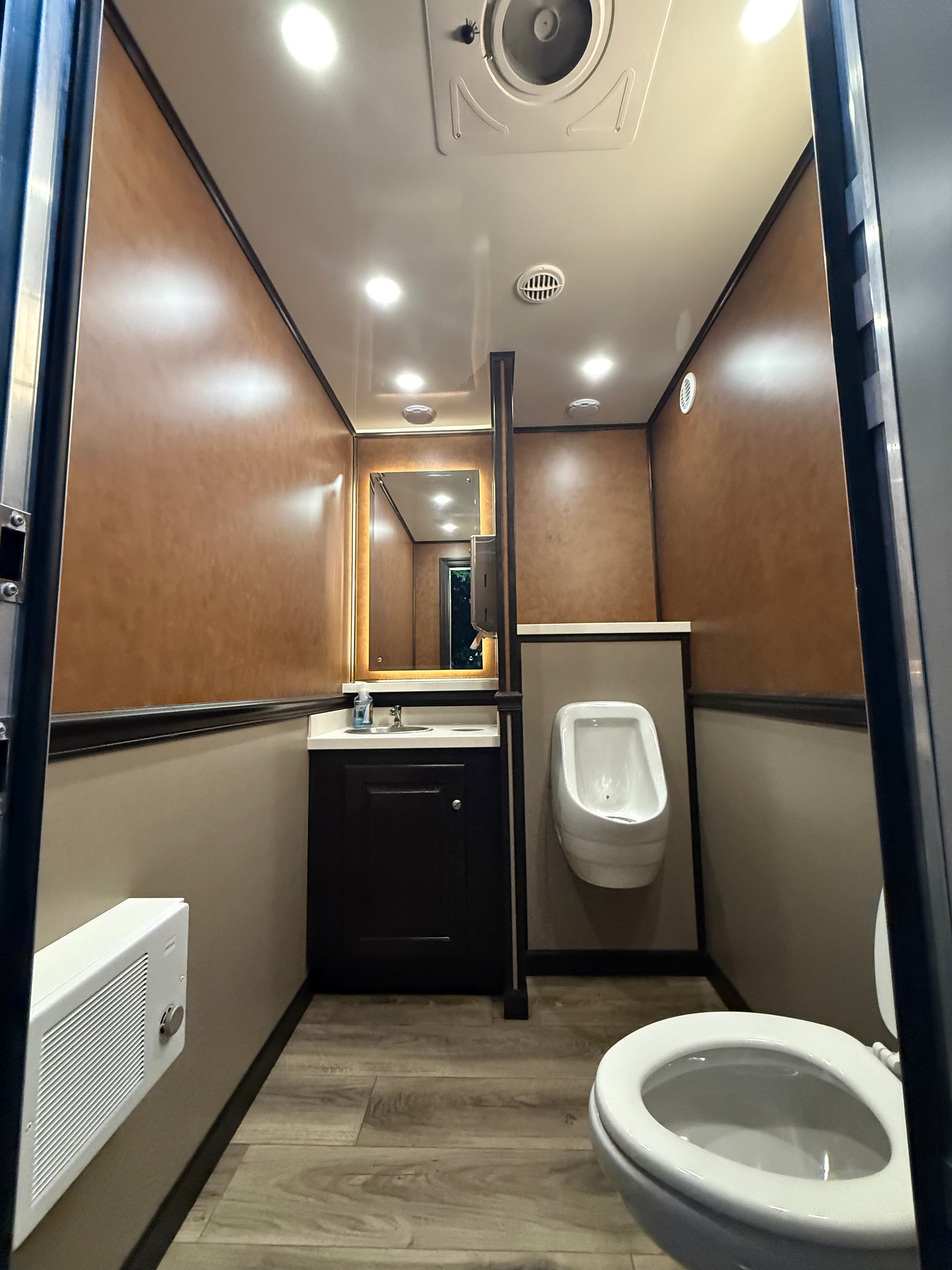 Luxury restroom trailer interior with toilet, urinal, vanity sink, and illuminated mirror, featuring upscale finishes and wood-look flooring for event rentals.