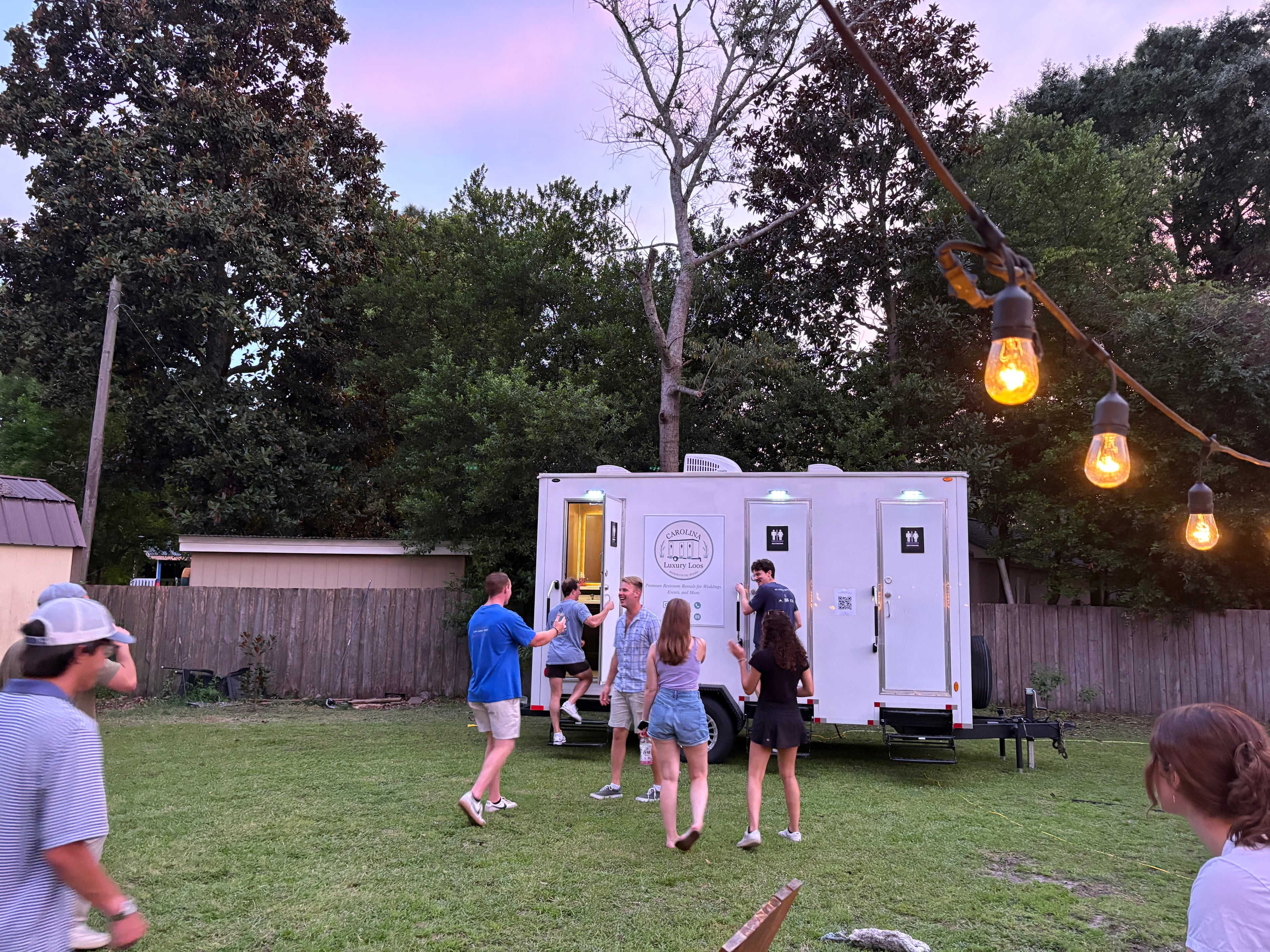 Backyard event with people near Carolina Luxury Loos’ 3‑stall luxury restroom trailer under string lights in Wilmington, NC.